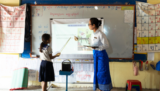 A young girl and teacher in a classroom pointing at text and images projected onto the board.